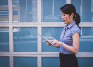 Photograph of an Asian business woman reading something on an ipad, in front of a set of office windows.